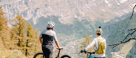 Two people on a mountain trail with bicycles, surrounded by snow-capped mountains and trees.