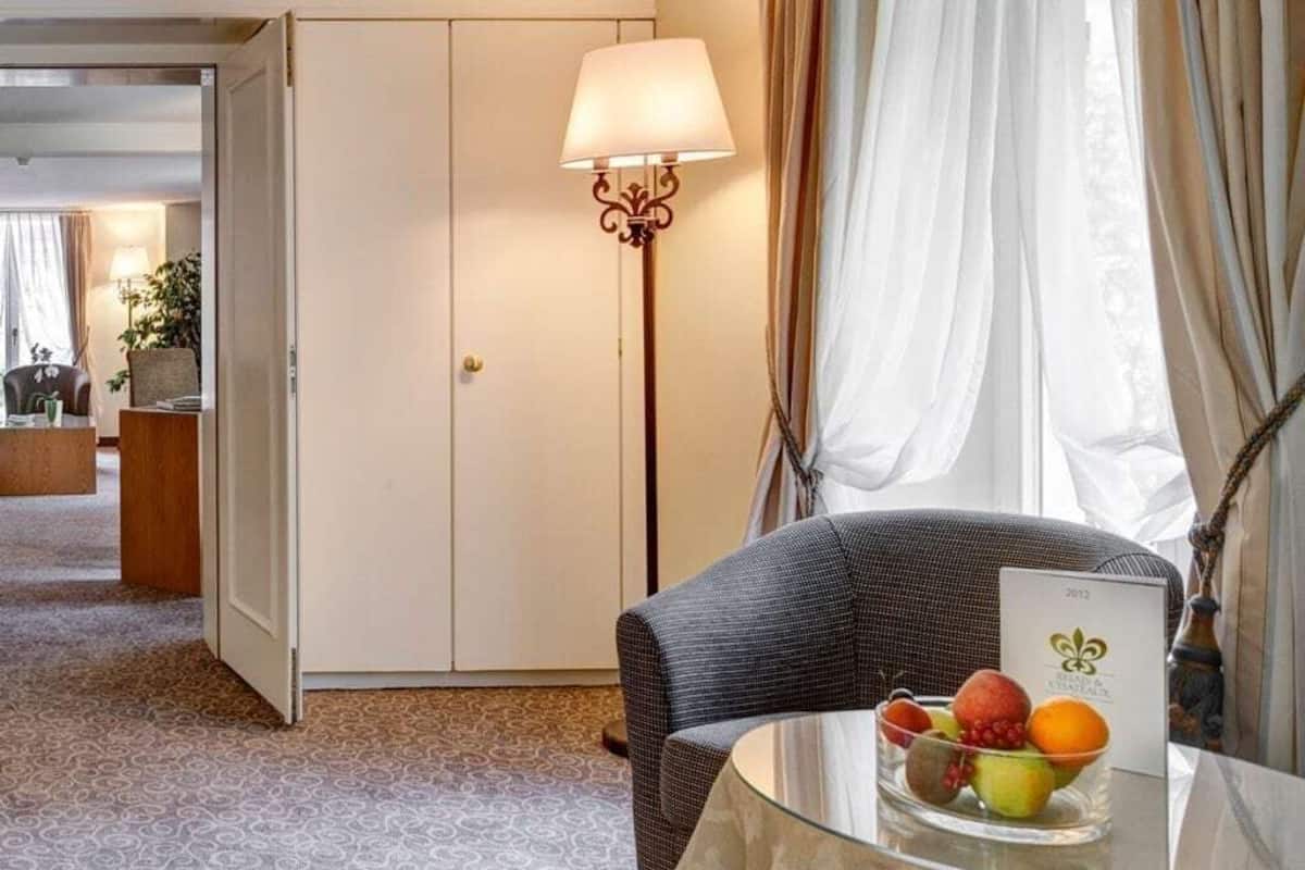 Une chambre d’hôtel avec un lit, un bureau sur lequel se trouve un panier de fruits, une lampe et une vue sur l’extérieur à travers des rideaux transparents.