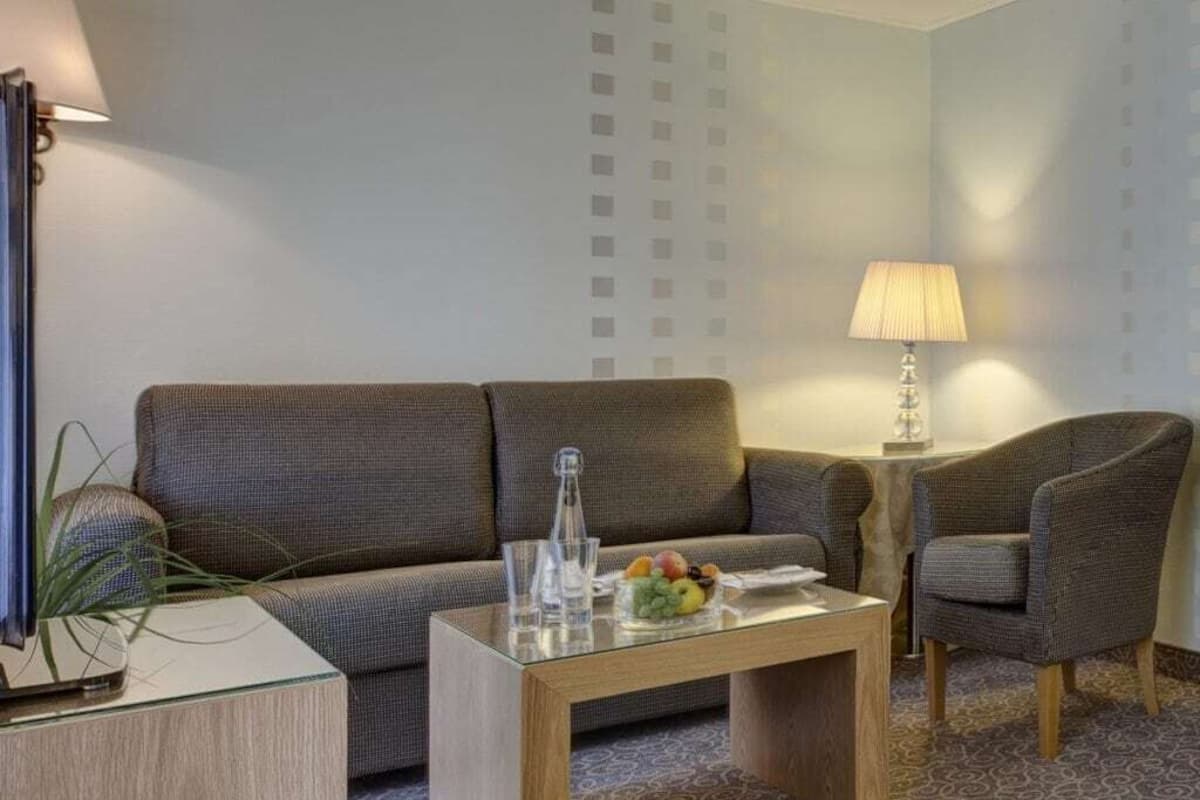 Une chambre d’hôtel moderne avec un canapé, une table basse en bois sur laquelle sont disposés des fruits, une lampe et une table d’appoint.