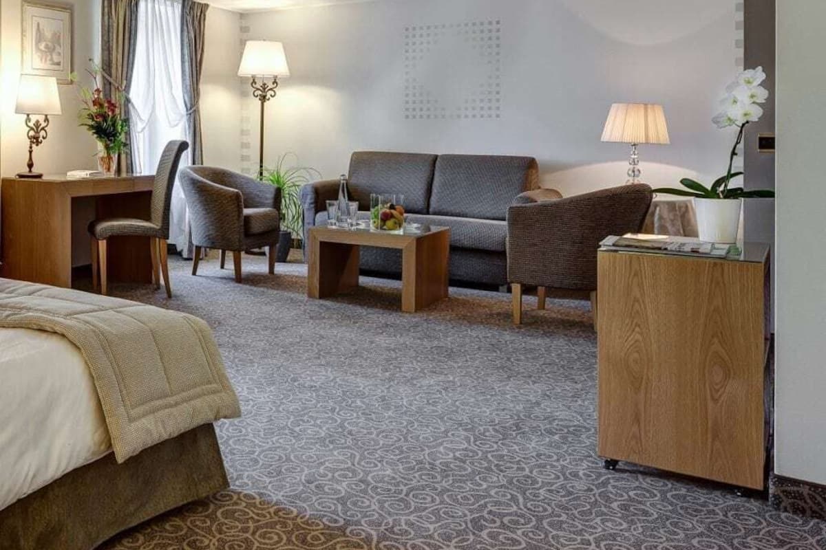 Une chambre d’hôtel comprenant un lit, un canapé, deux fauteuils, une table basse, un bureau avec une lampe, et une petite table d’appoint avec une lampe et un vase de fleurs.