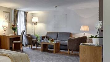 Ein Hotelzimmer mit einem Bett, einer Couch, zwei Sesseln, einem Couchtisch, einem Schreibtisch mit Lampe und einem kleinen Beistelltisch mit Lampe und einer Blume.