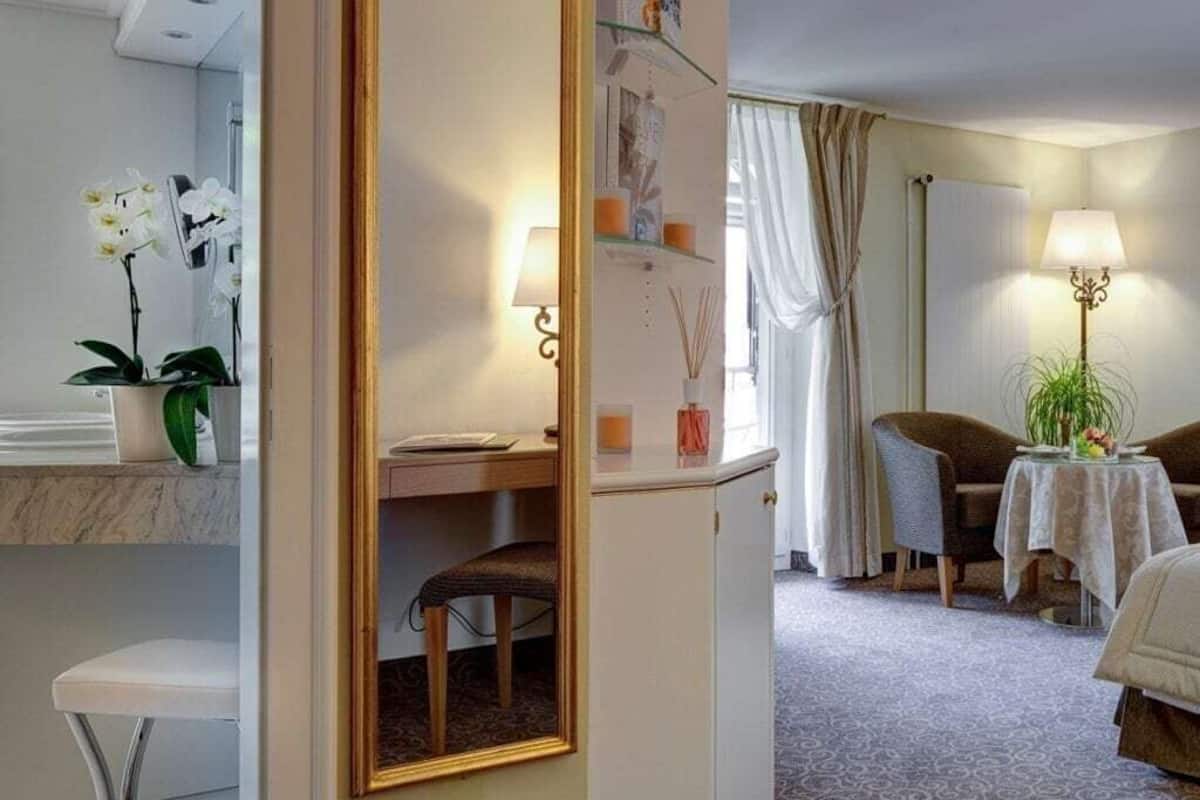 Une chambre d’hôtel comprenant un lit, un bureau avec une chaise, une petite table avec une lampe, un miroir et une plante en pot.