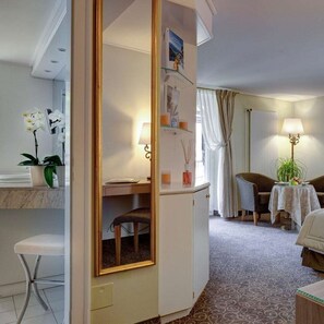 A hotel room with a bed, a desk with a chair, a small table with a lamp, a mirror, and a potted plant.
