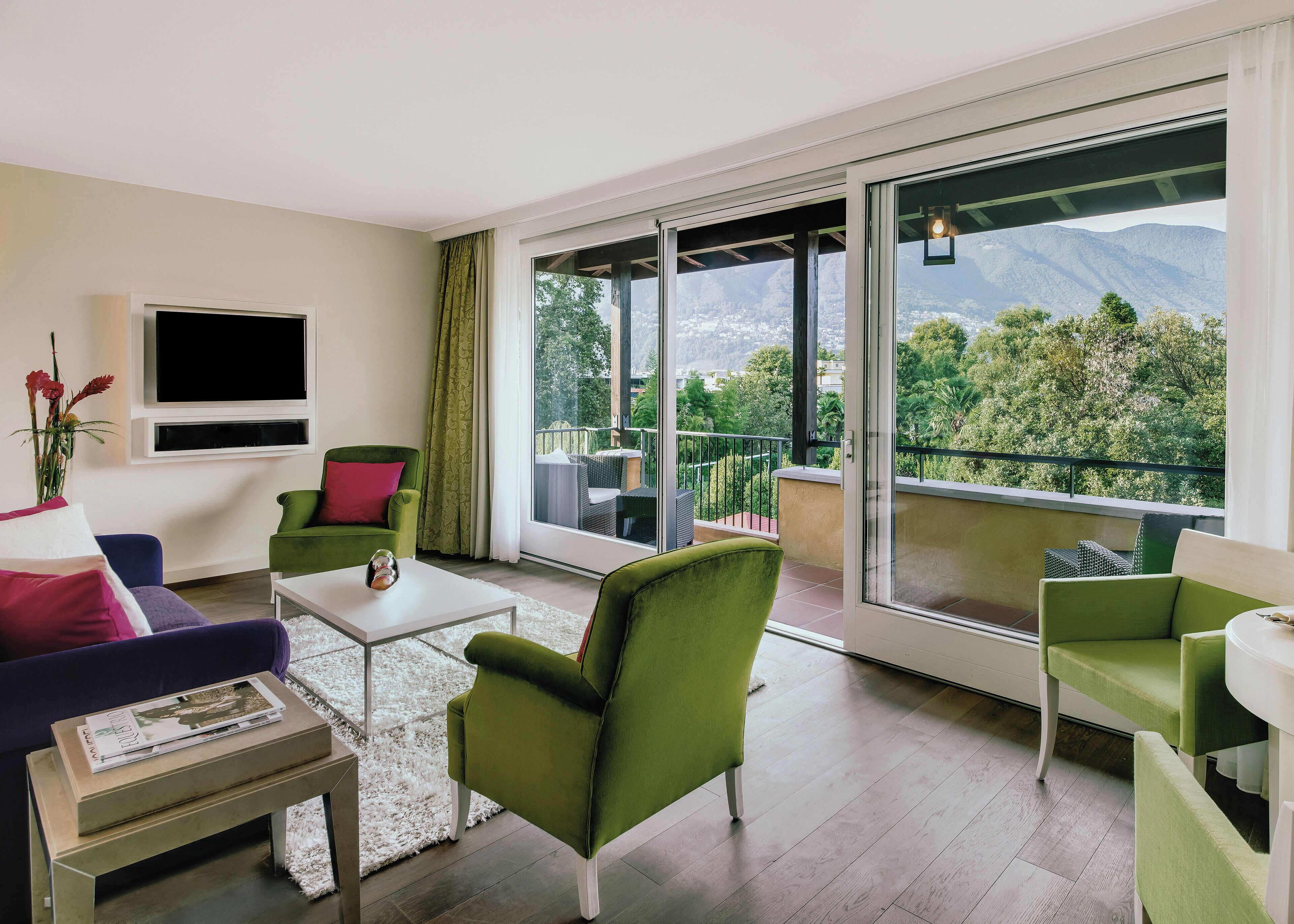 Un salon moderne avec des meubles verts et violets, un téléviseur à écran plat et un balcon offrant une vue sur les montagnes.