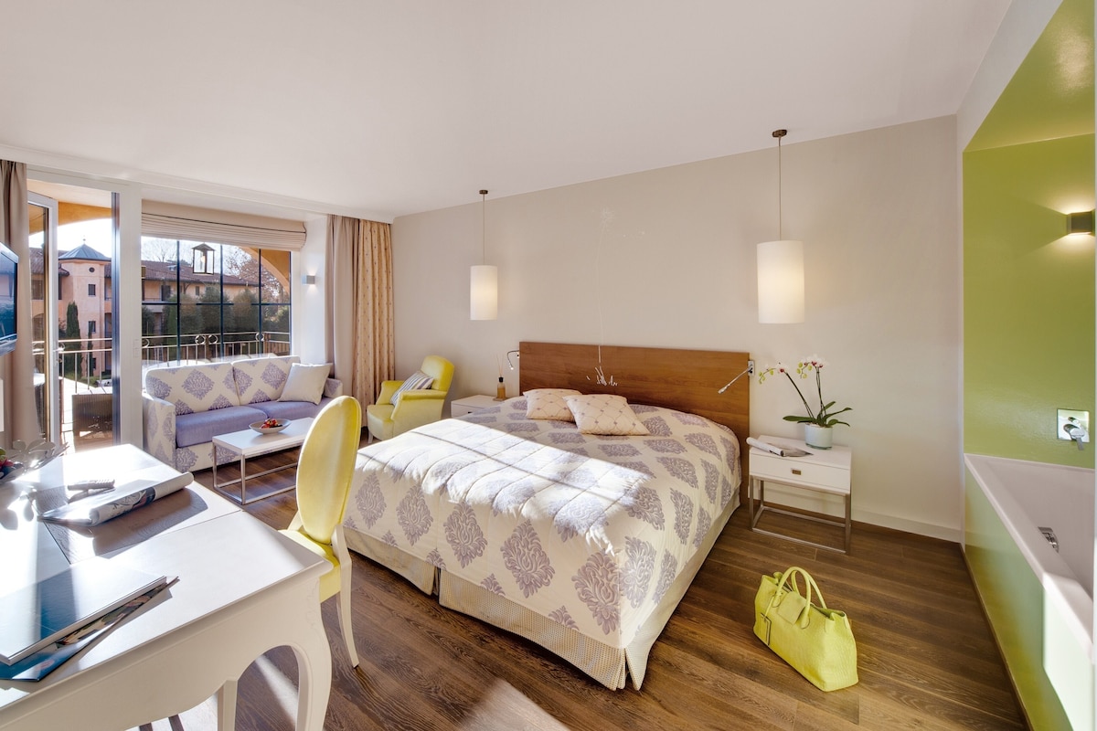 Habitación de hotel con cama, sofá, escritorio y silla. Se ve una ventana con cortinas, una mesita de noche con lámpara y una planta en maceta.