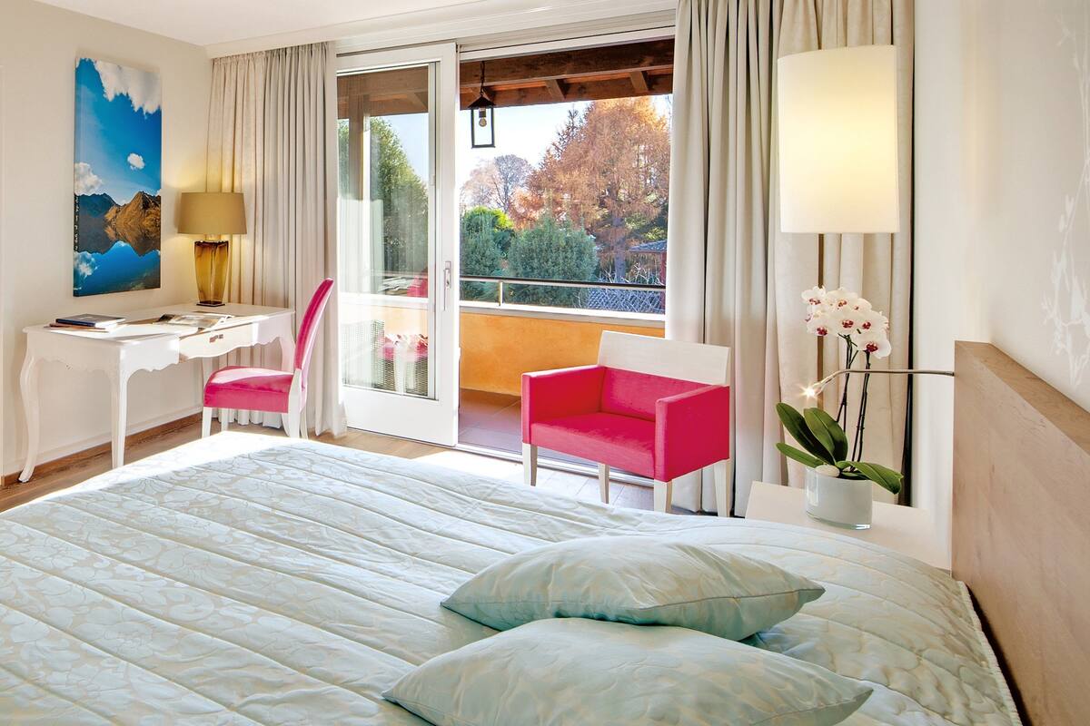 A hotel room with a large bed, a desk, a chair, and a balcony with a view of trees.