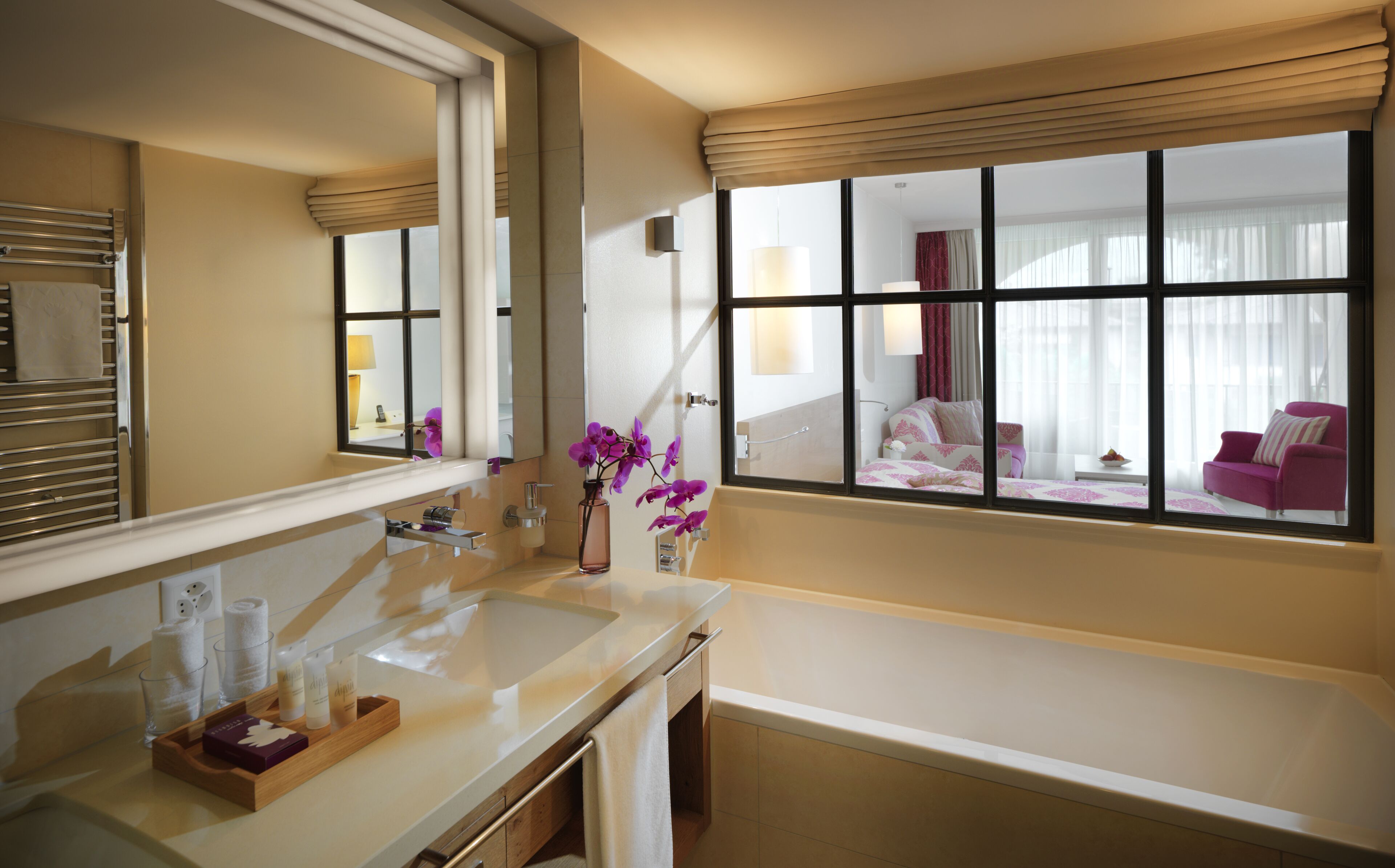 A hotel bathroom with a large mirror, a sink, and a window overlooking a bedroom.