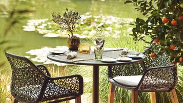 A round table with a potted plant, a glass, and a bottle, surrounded by wicker chairs, set against a pond with lily pads and greenery.