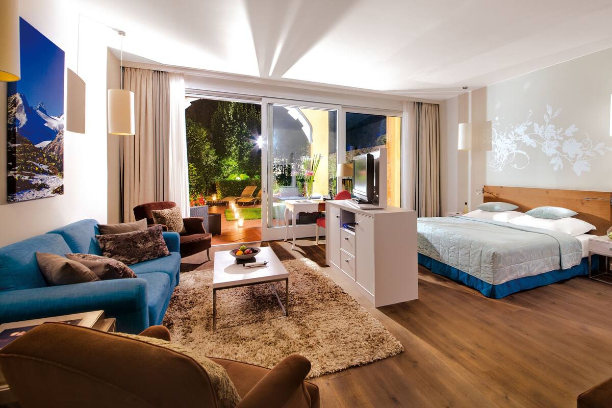 A modern hotel room with a large bed, a desk with a computer, a blue sofa, and a view of the garden through the glass door.
