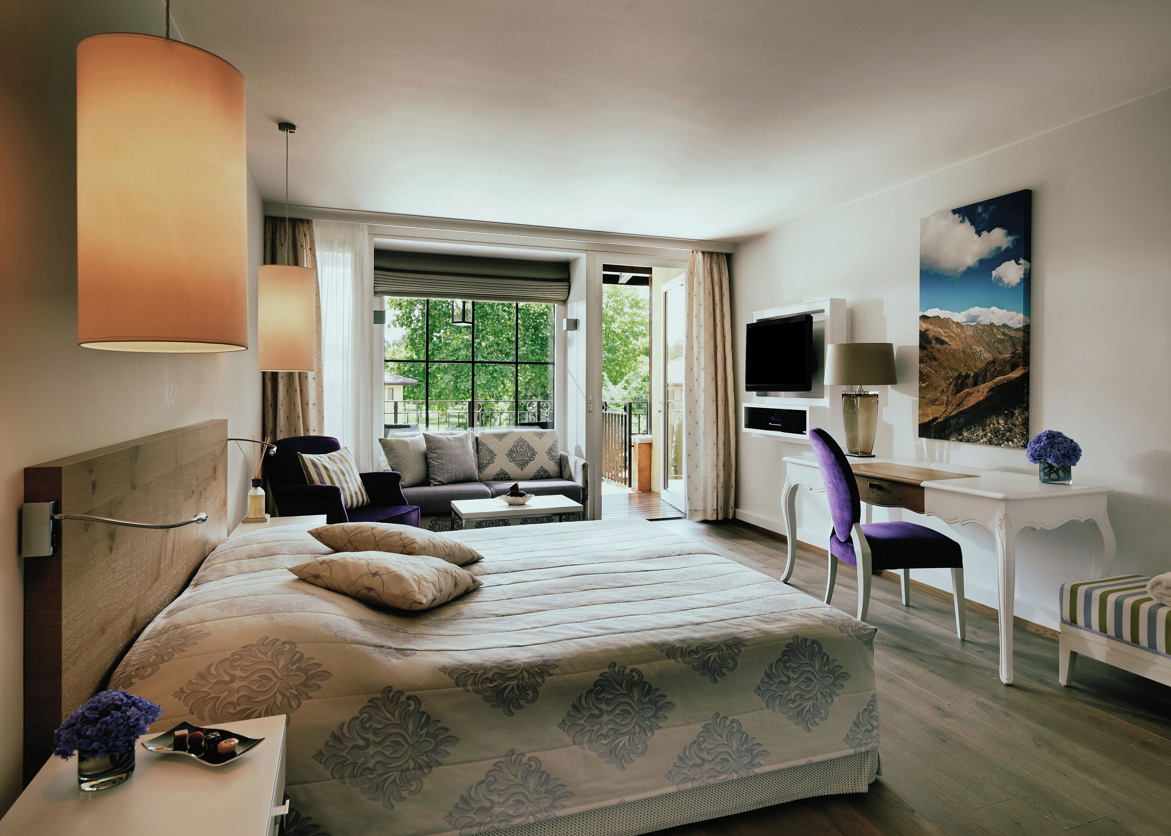 Habitación de hotel con cama, escritorio, silla, sofá, televisor y ventana con vista a follaje.