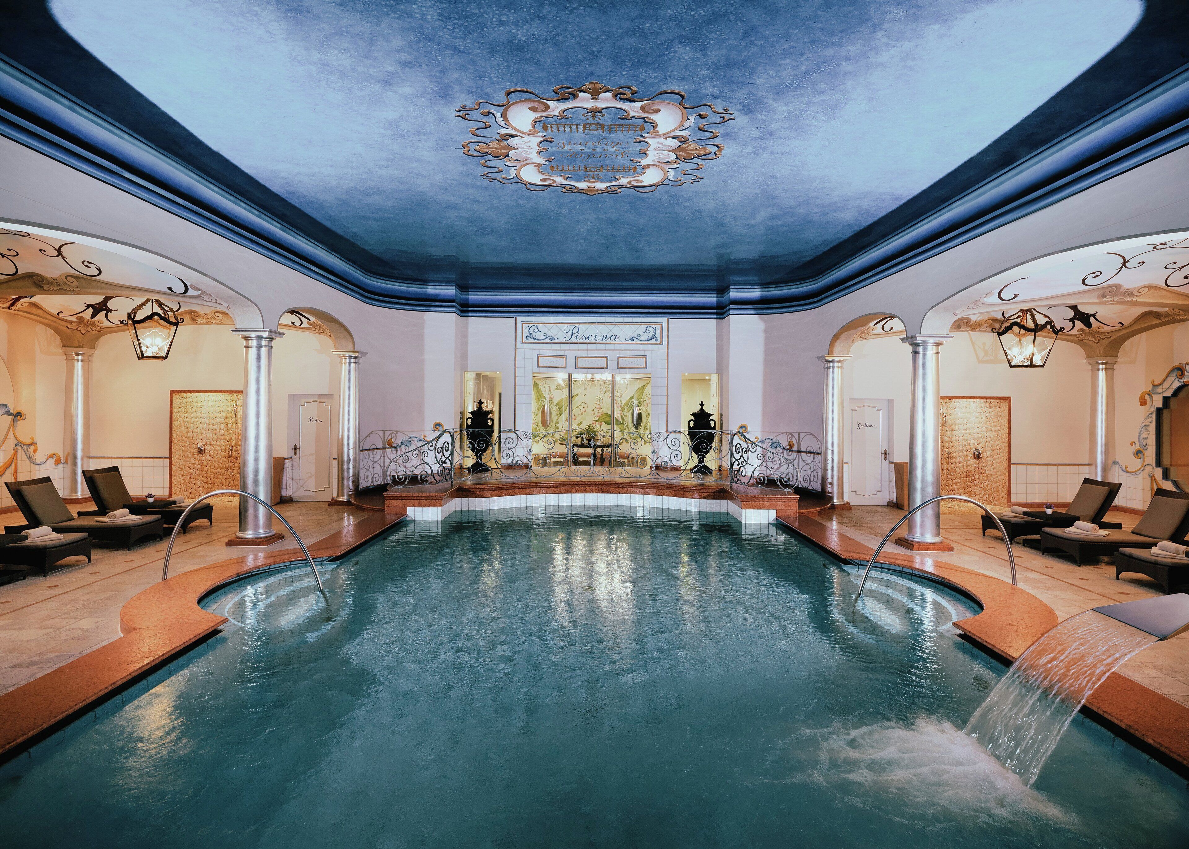 Un área de piscina interior lujosa con una fuente central, asientos elegantes y ornamentales decoraciones en el techo.