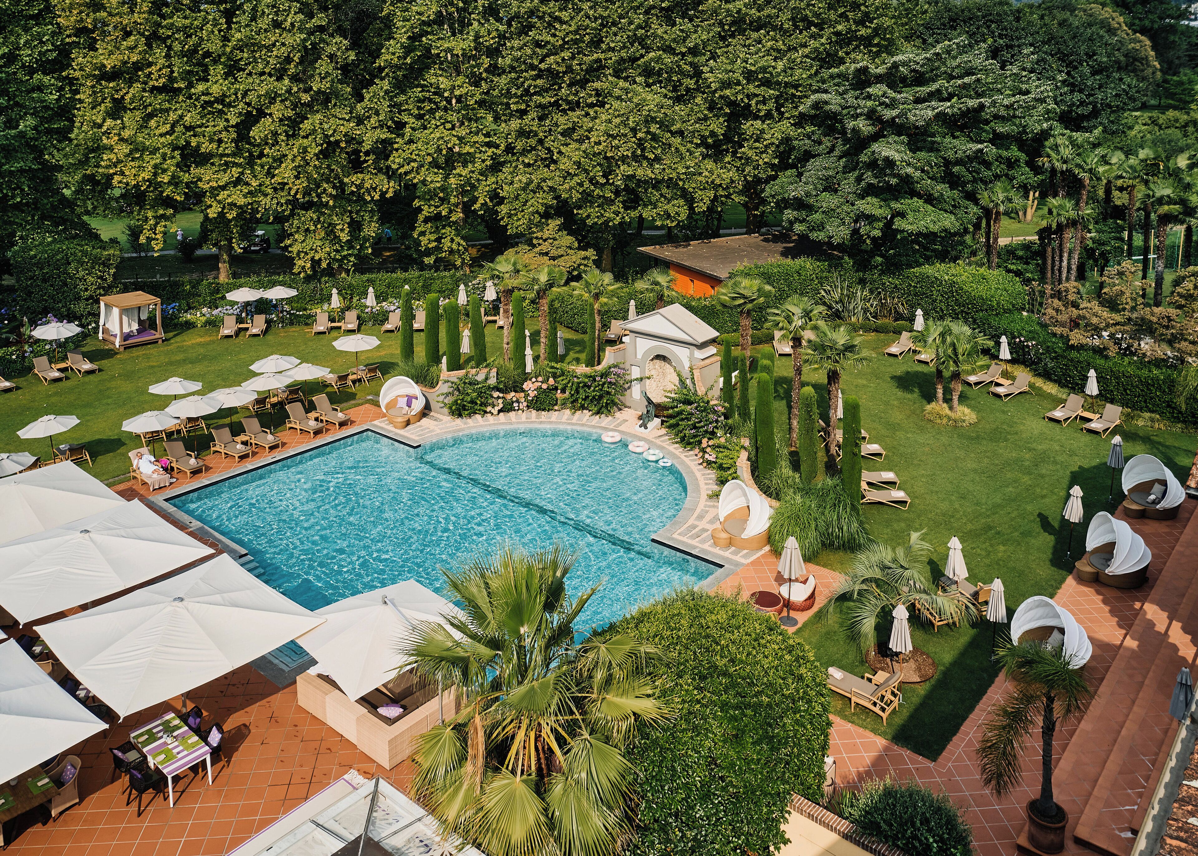 Una gran piscina rodeada de sillas de descanso y sombrillas, con un jardín cuidado y árboles al fondo.