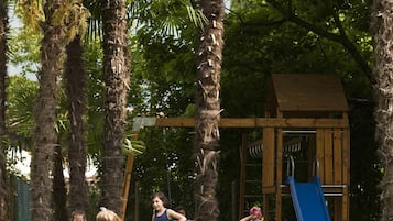Children can play on the playground's slide, swing set, and climbing frame amidst lush greenery and tall palm trees.