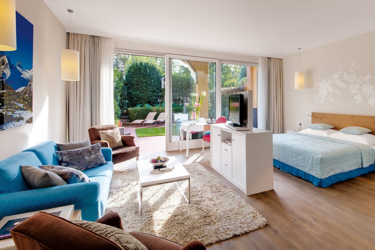 A modern hotel room with a large bed, a sofa, a desk with a TV, and a view of a garden through the sliding glass door.