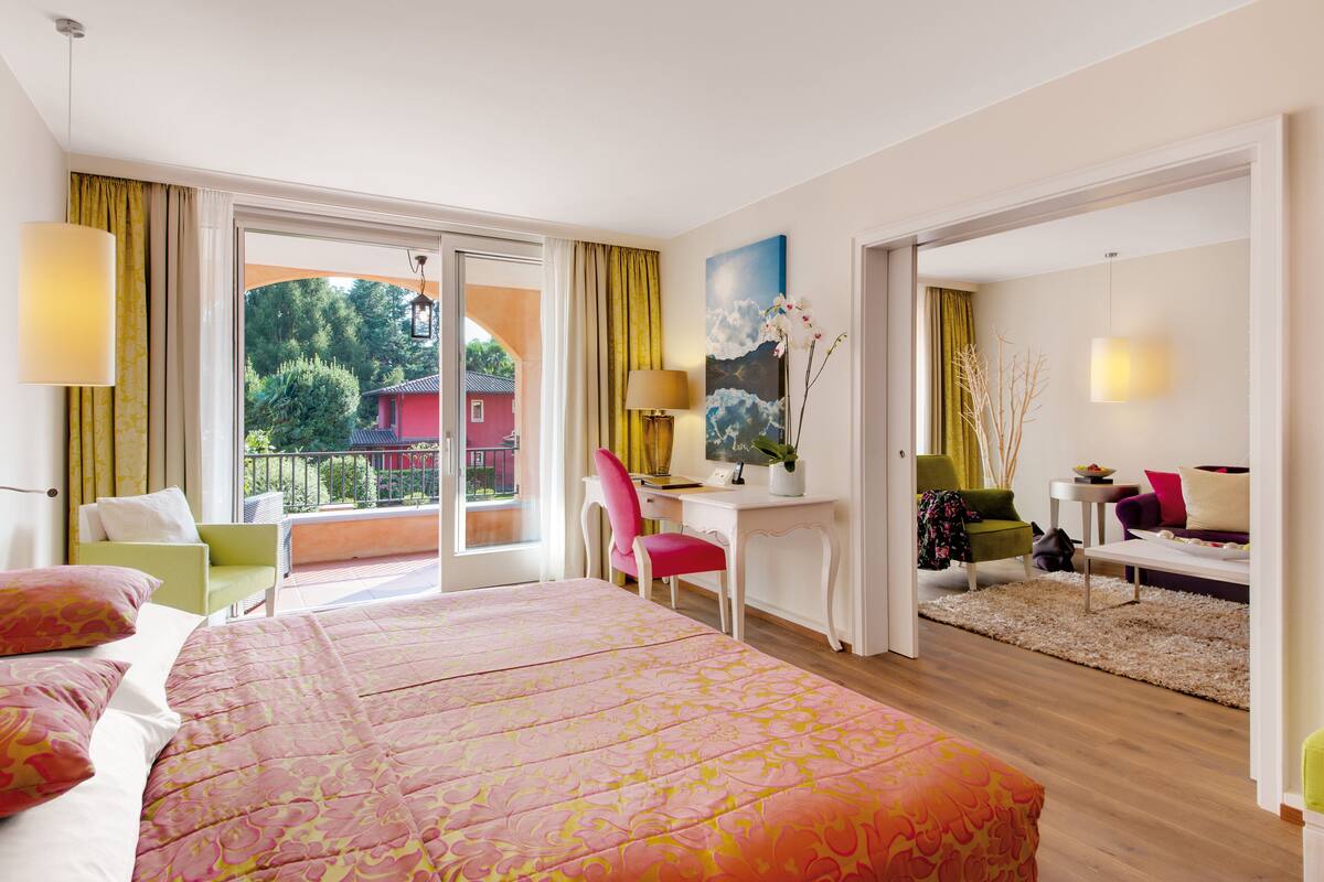 A hotel room with a large bed, a desk, a chair, and a balcony with a view of trees and a building.
