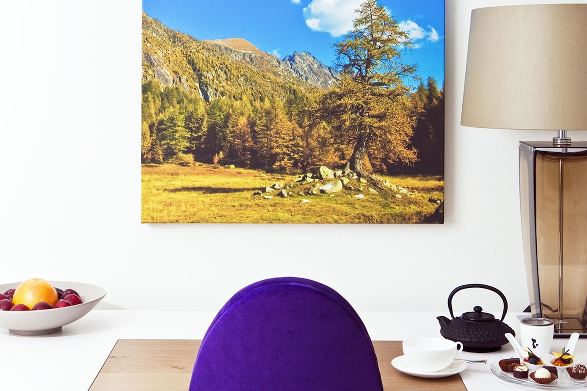 Una habitación con un cuadro de gran formato que muestra un paisaje, un escritorio de madera, una silla morada, una mesa blanca, una lámpara, un calentador de agua y una bandeja con postres.