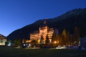 Un gran hotel de varios pisos con una torre destacada y una bandera, rodeado de un patio bien iluminado y árboles.