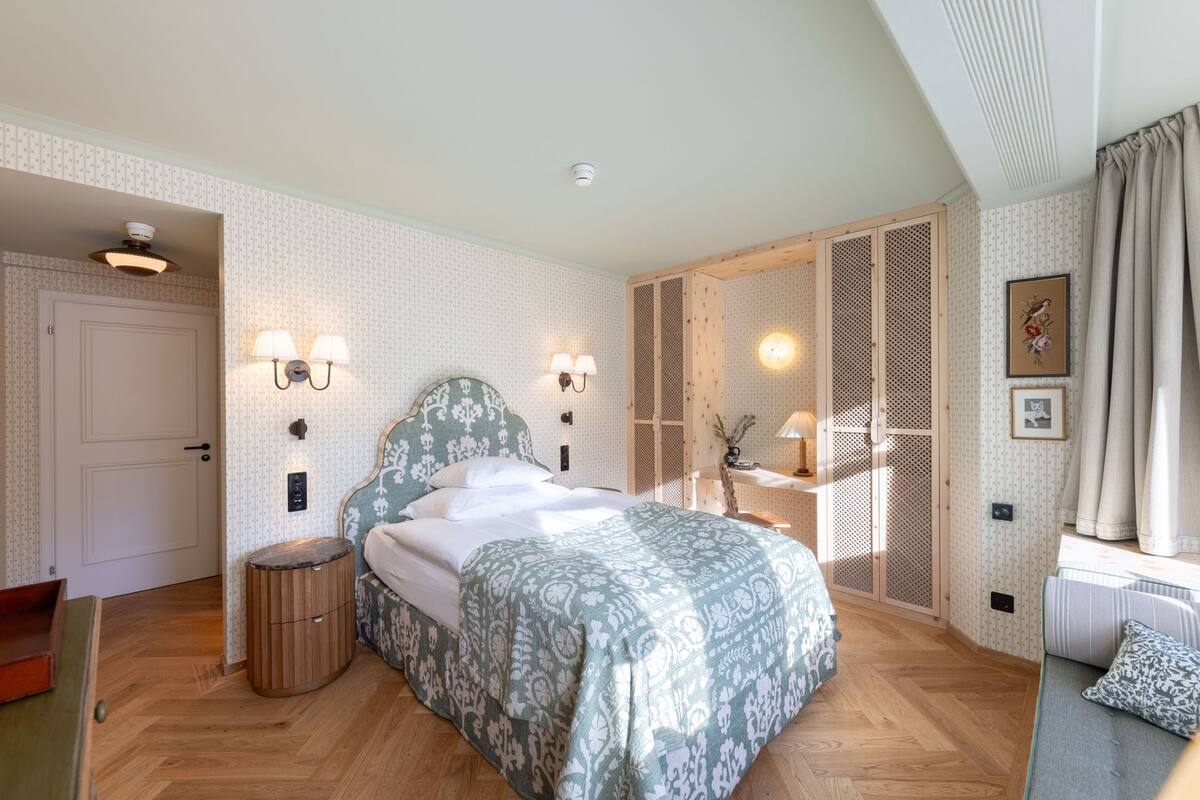 Ein Schlafzimmer mit Bett, Nachttischlampen, Holzboden, Kleiderschrank und einem Fenster mit Vorhängen.