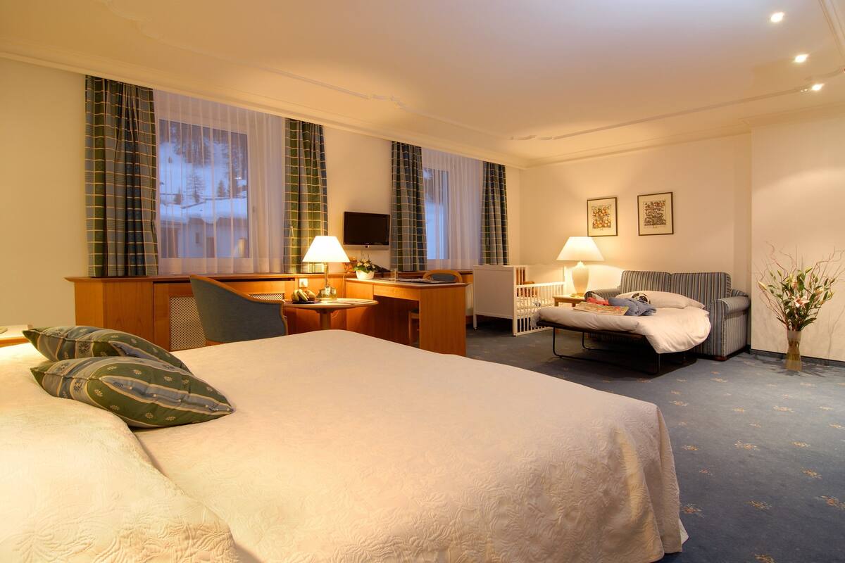 Ein Hotelzimmer mit einem großen Bett, einem Schreibtisch mit Stuhl, einem kleinen Tisch, einem Sessel, einem Fernseher und einem Fenster mit Vorhängen.