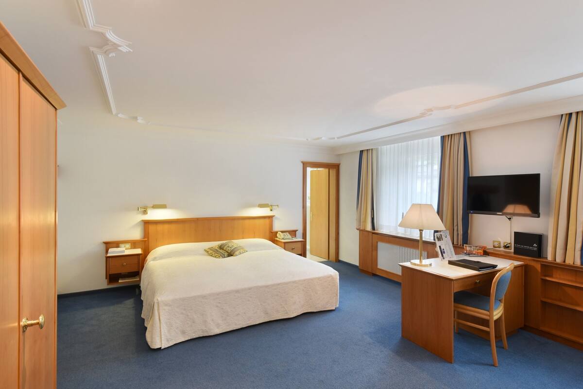 Ein Hotelzimmer mit einem großen Bett, einem Schreibtisch mit Stuhl, einem Fernseher und einem Fenster mit Vorhängen.