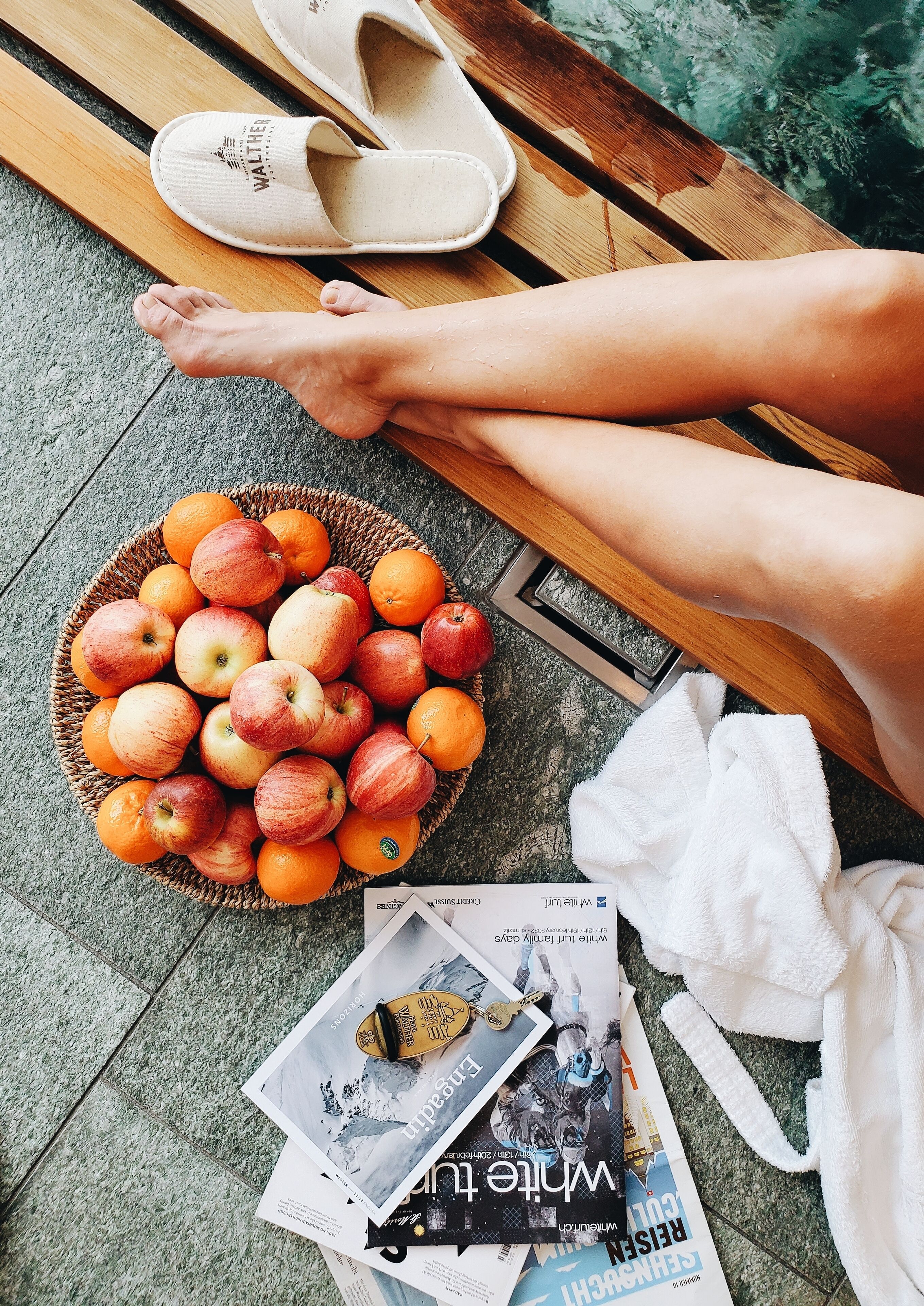 A wooden bench with a pair of white slippers, a basket of apples and oranges, and some magazines on a tiled floor.