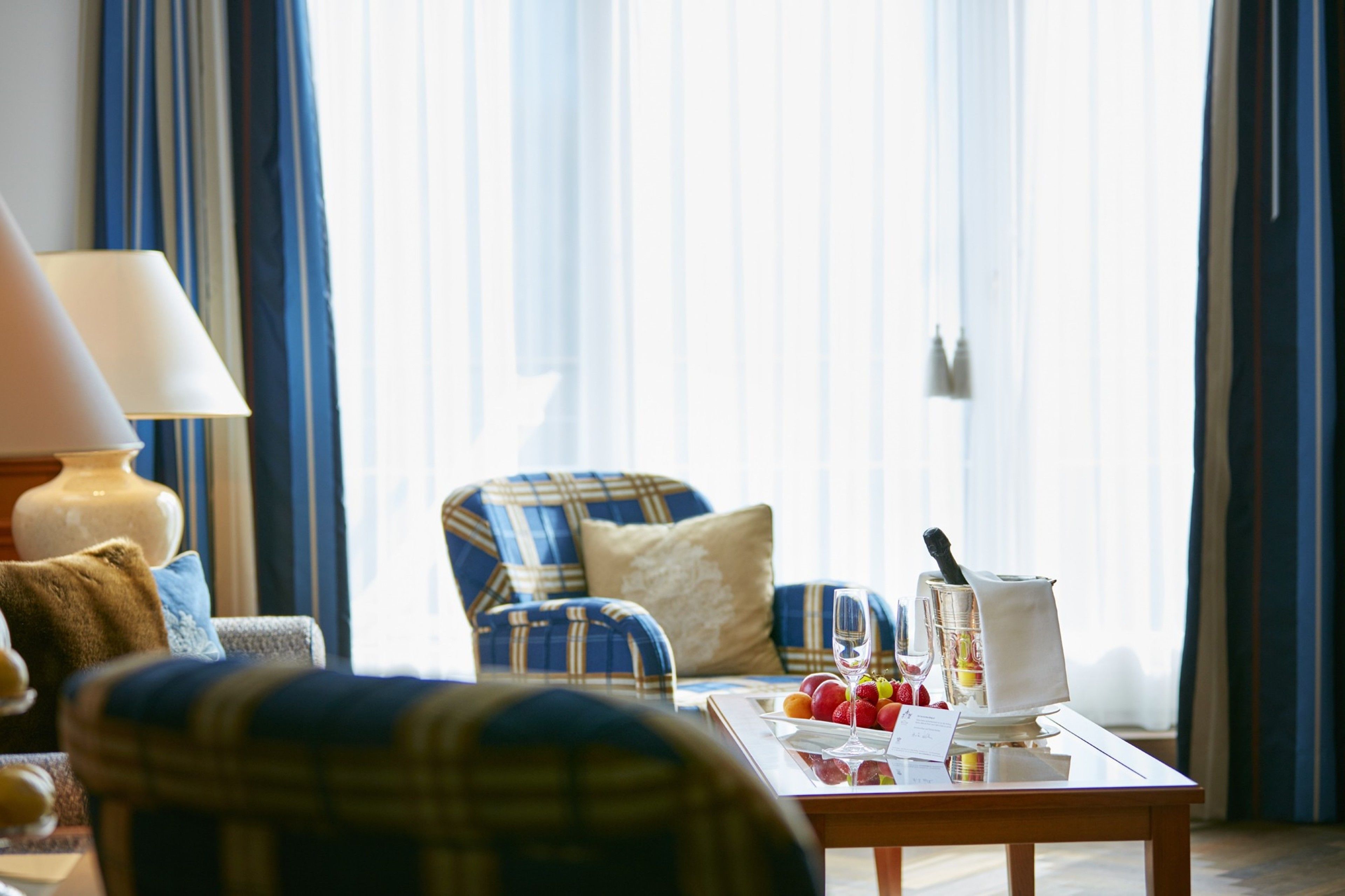A hotel room with a plaid armchair, a table with fruit and a bottle, and a lamp.