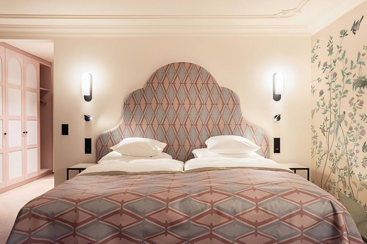 Ein Schlafzimmer mit einem gemusterten Bettbezug, einem dekorativen Kopfteil, Wandleuchten und floralem Wandpapier auf der rechten Seite.
