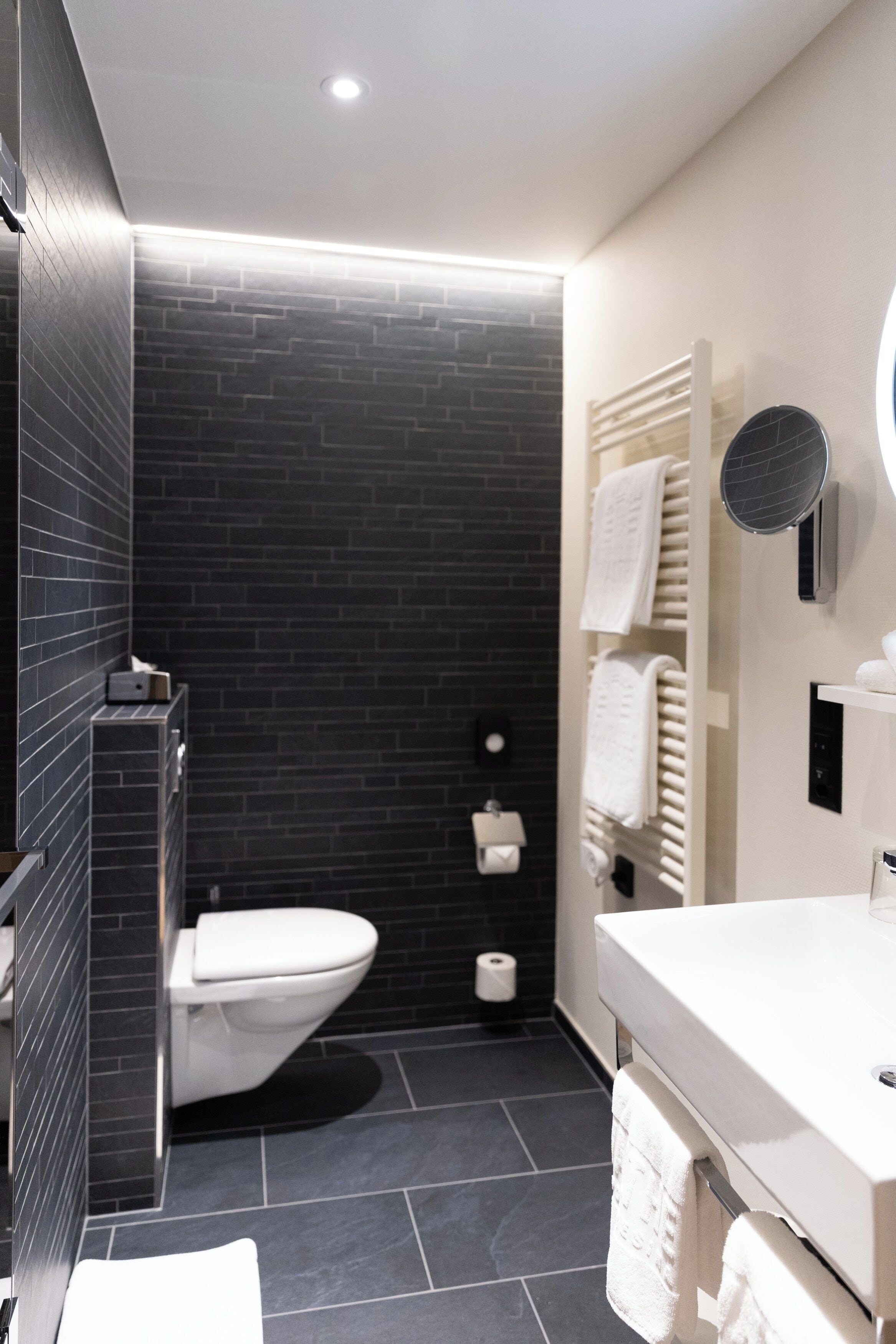 A modern bathroom with a black tiled wall, a white toilet, a white sink, and a towel rack.