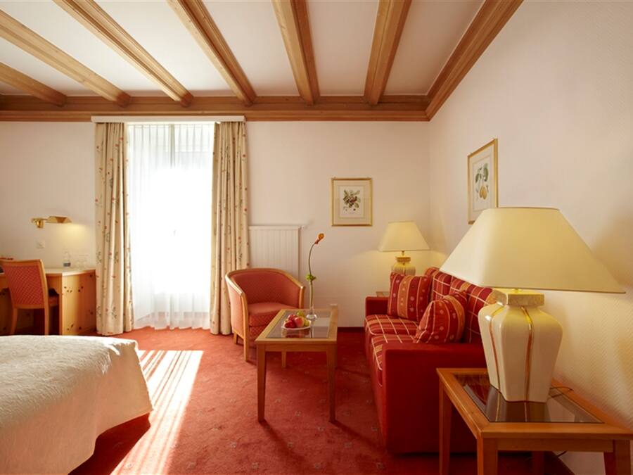 Habitación de hotel con cama, sofá rojo, mesa de centro de madera, escritorio con silla, dos lámparas y ventana con cortinas.