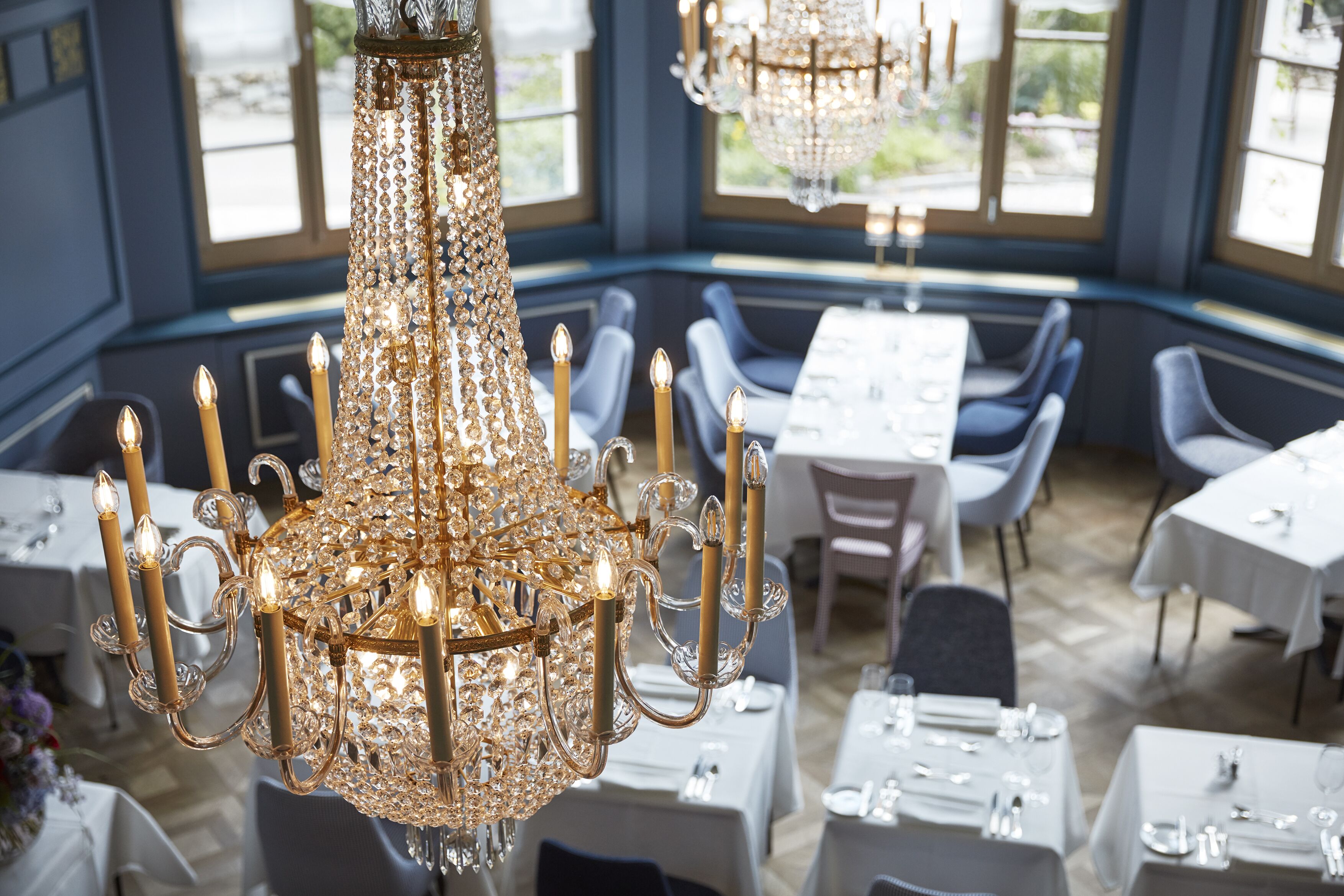 A chandelier with multiple candle-like lights, tables with white tablecloths, and blue chairs in a dining area with large windows.