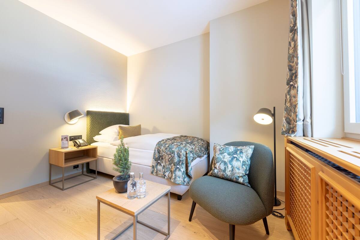 Ein modernes Hotelzimmer mit Bett, Sessel, kleinem Tisch, Nachttisch, Lampe und einem Fenster mit Vorhängen.