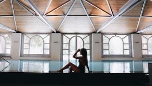 A person sitting by a pool with a wooden ceiling and large arched windows. - Hotel Walther (Pontresina)