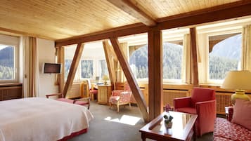 A room with a bed, a sofa, a chair, a table, a TV, and large windows with a mountain view.
