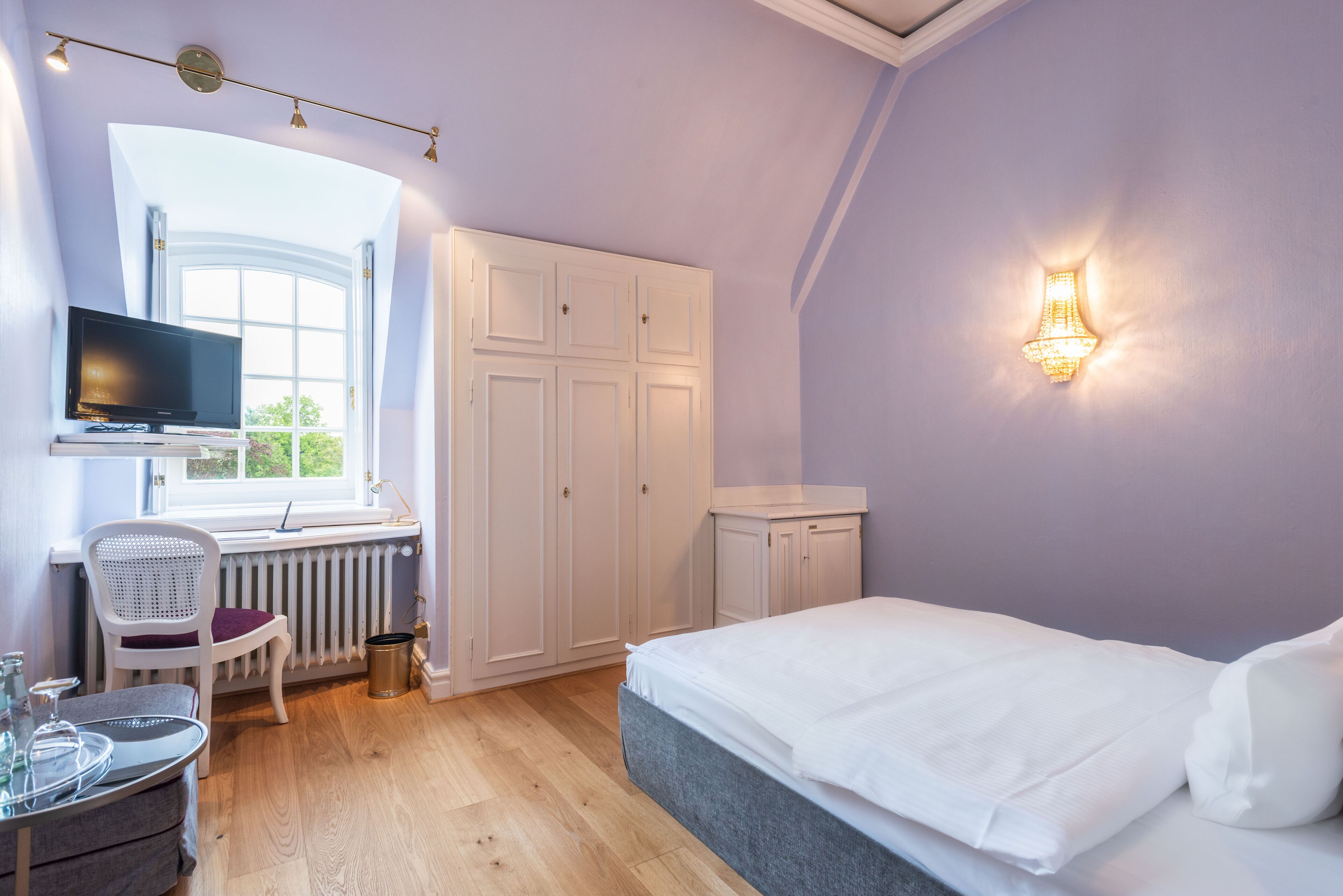 A bedroom with a bed, a TV, a desk with a chair, and a window.