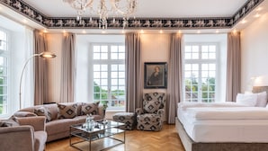 Une pièce spacieuse comprenant un grand lit, un canapé, une table basse et un fauteuil. De grandes fenêtres sont agrémentées de rideaux, et la pièce est décorée d’un lustre et d’un tableau encadré accroché au mur.