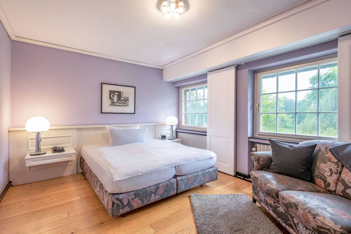 Une chambre à coucher avec un lit, des tables de chevet, un canapé, une fenêtre et un tableau encadré au mur.