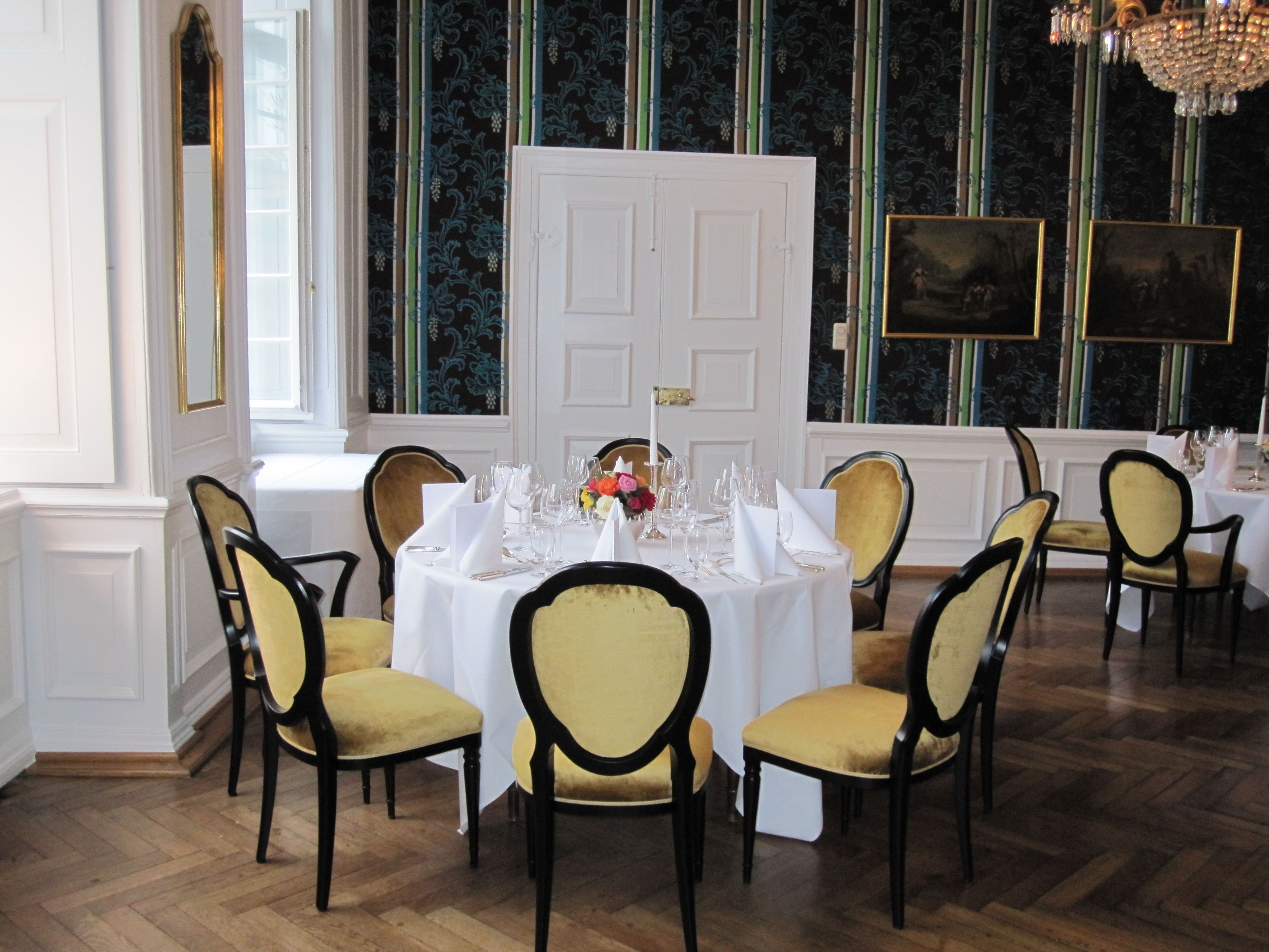 Ein formeller Speisesaal mit einem runden Tisch, gedeckt für sechs Personen, umgeben von sechs Stühlen mit hohen Rückenlehnen und gepolsterten Sitzflächen. Der Raum verfügt über eine große, weiße Tür, ein Fenster mit Blick ins Freie und eine Wand mit gerahmten Kunstwerken.