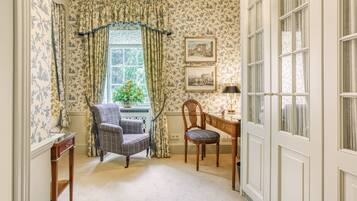 A room with floral wallpaper, a window with curtains, a chair, a desk, and a lamp.