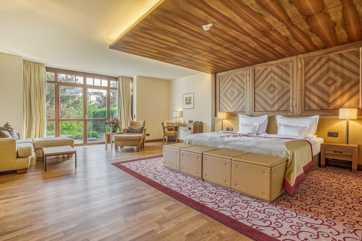 Ein geräumiges Schlafzimmer mit einem großen Bett, einem Holzkopfteil und Blick auf Grün hinter großen Fenstern.