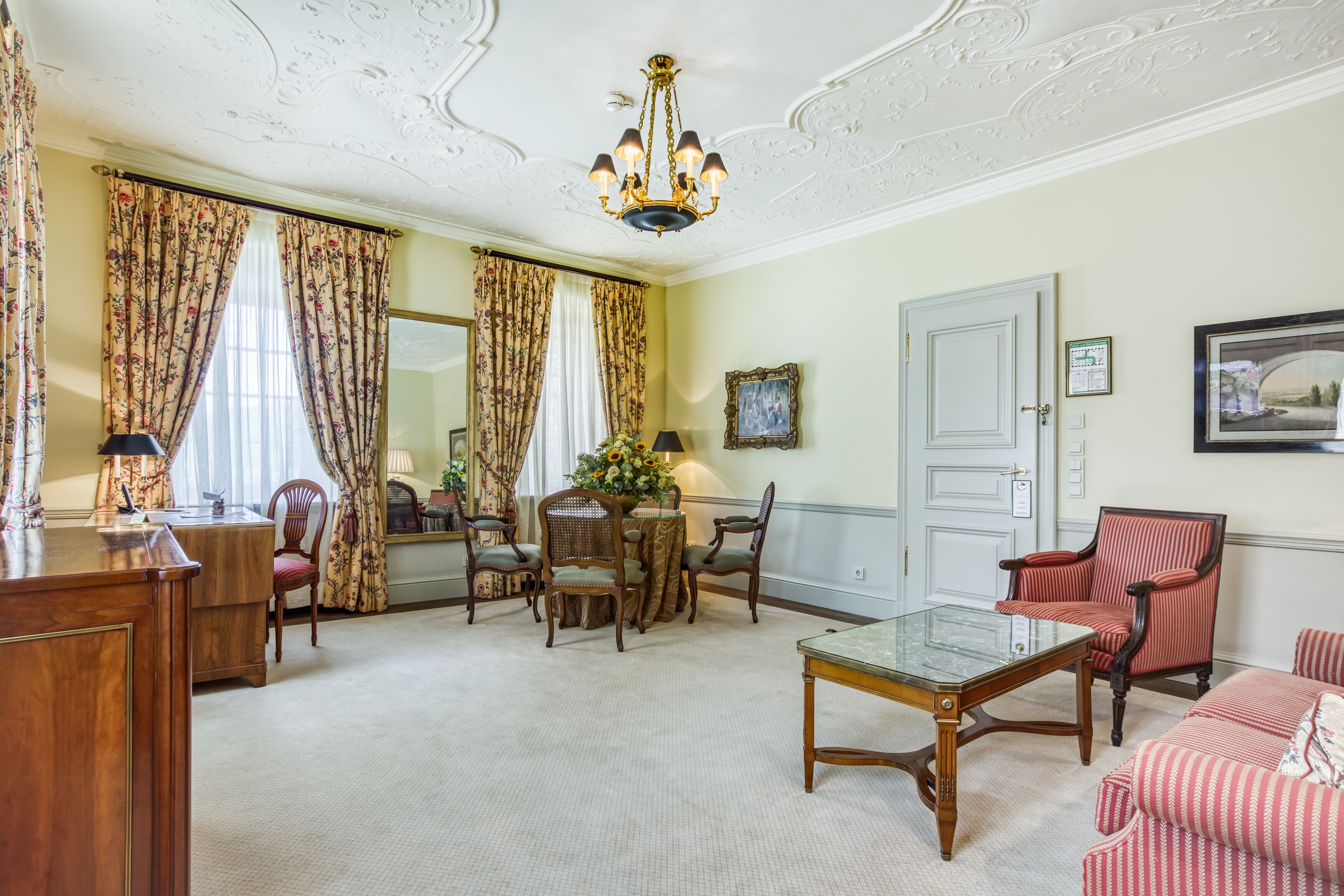 A well-furnished living room with a wooden coffee table, a red striped armchair, and a dining area with floral curtains and a flower vase.