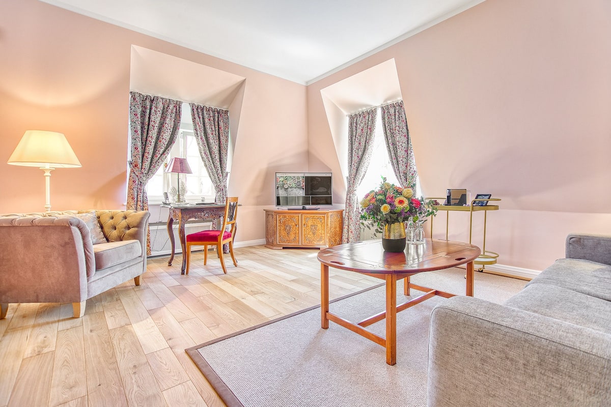 Ein Wohnzimmer mit Sofa, Sessel, Couchtisch und Fernseher. Es gibt ein Sideboard, eine Lampe und eine Vase mit Blumen.