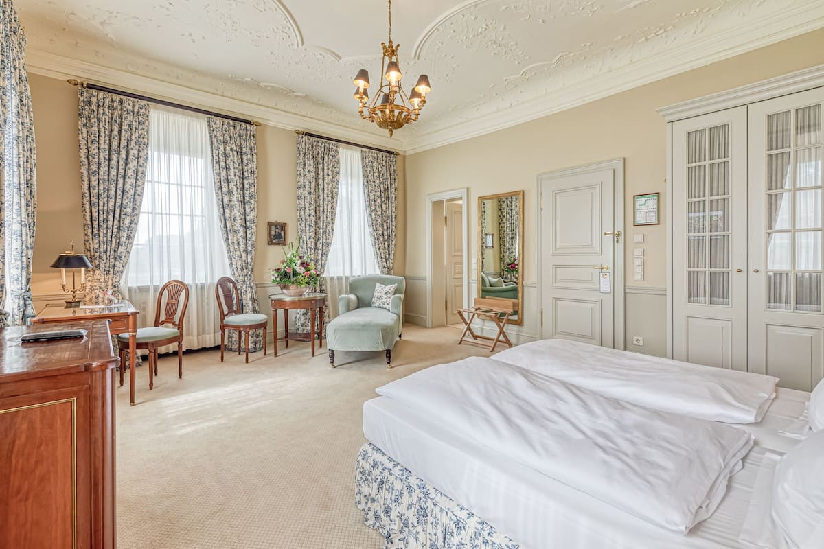 Ein geräumiges Hotelzimmer mit einem großen Bett, einem Schreibtisch, zwei Stühlen, einem kleinen Tisch mit Lampe und einem Kronleuchter.