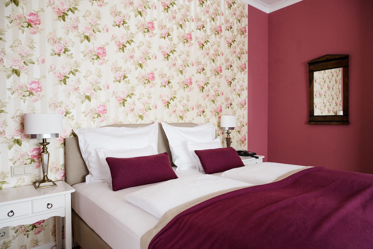 Ein Schlafzimmer mit floralem Wandpapier, einem Bett mit weißen Leinen und violetten Kissen, einem Nachttisch mit Lampe und einem Spiegel an der Wand.