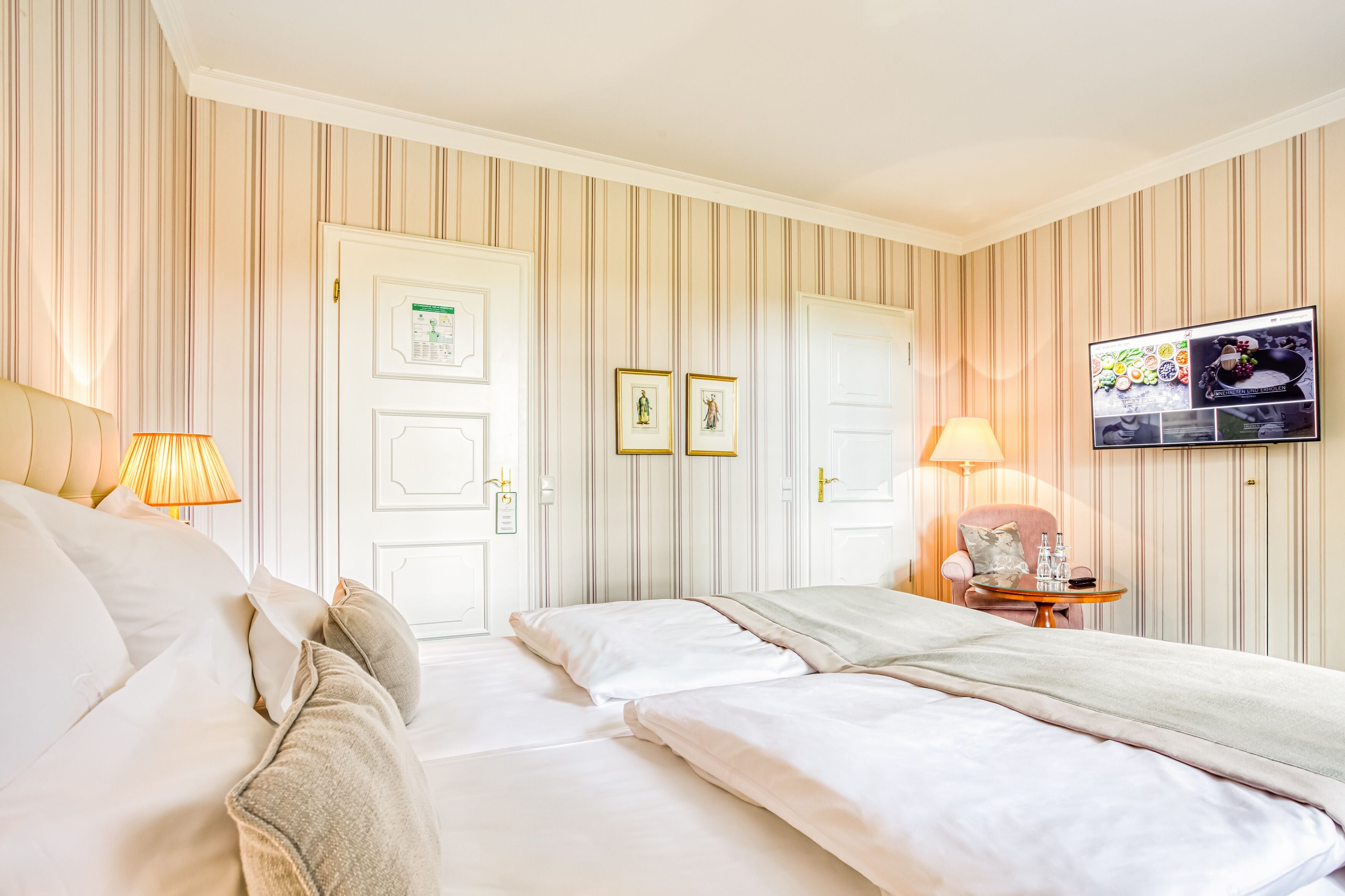 A hotel room with two beds, a TV, a desk with a lamp, and striped wallpaper.