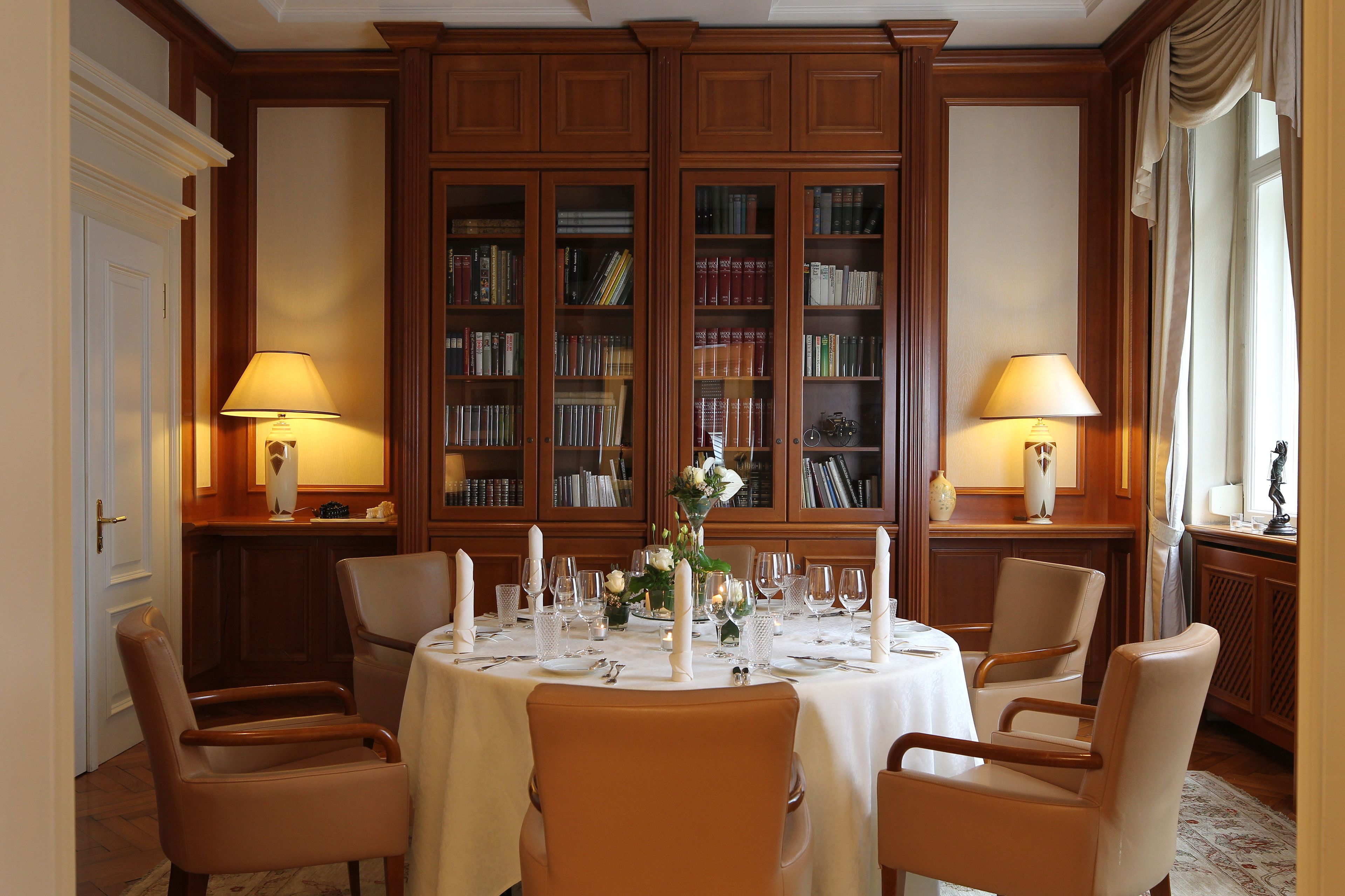 Un comedor con una mesa redonda dispuesta para cuatro personas, rodeada de sillas de madera. Se observan dos estanterías con libros detrás de la mesa, y dos lámparas a cada lado.