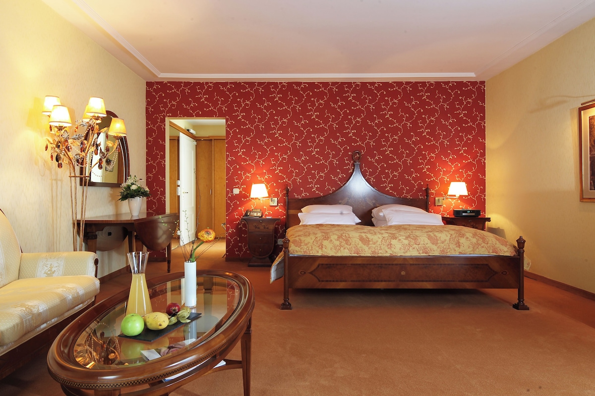 A bedroom with a bed, bedside tables, a mirror, a sofa, and a glass coffee table with fruits.