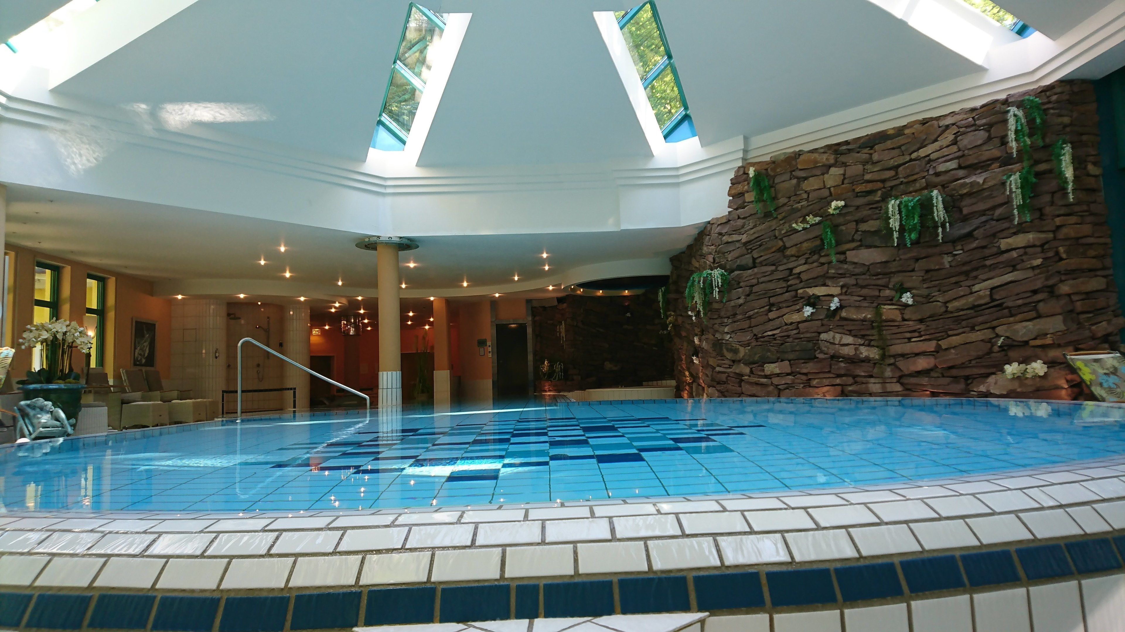 An indoor swimming pool with a tiled floor, a stone wall with greenery, and a skylight.