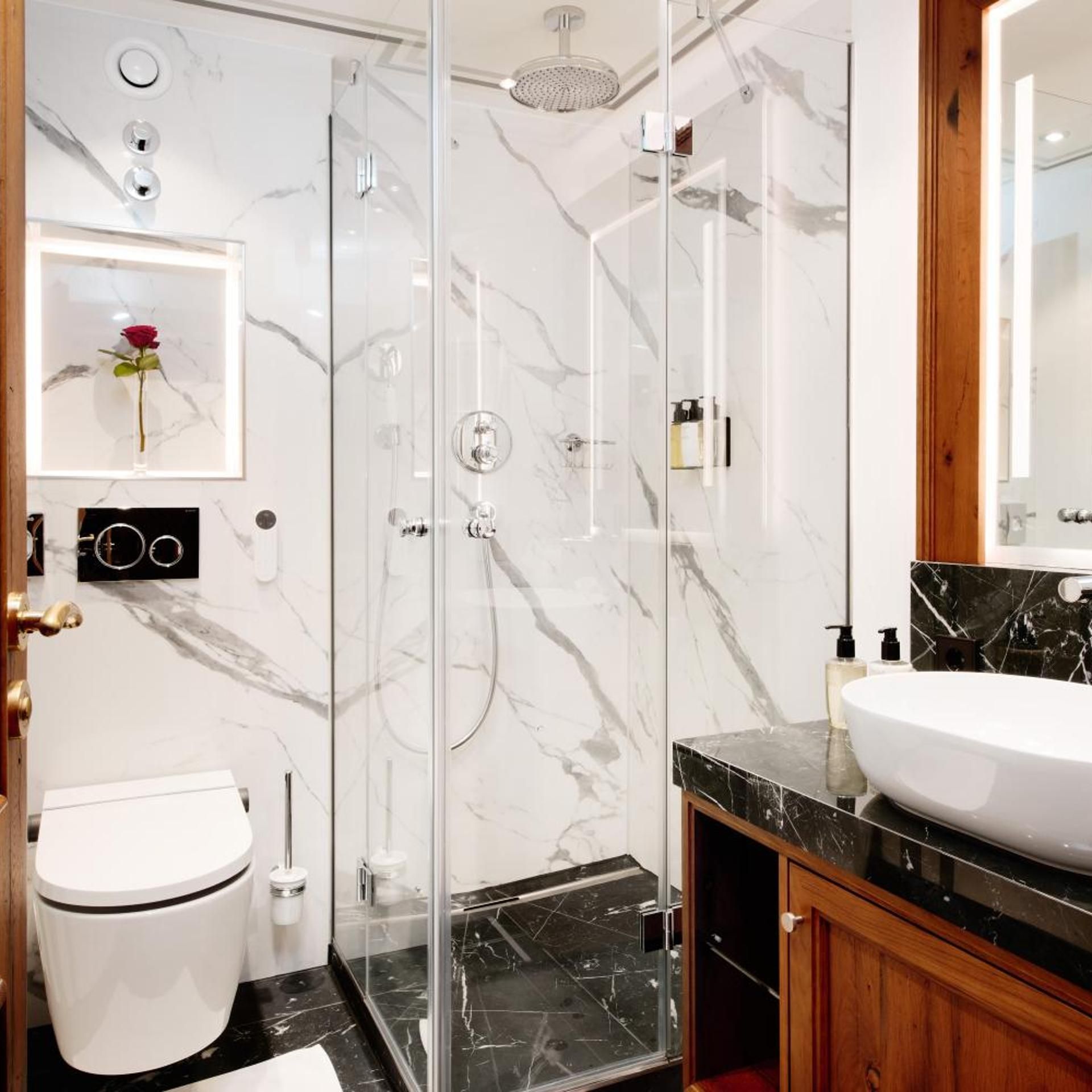 A modern bathroom with a glass-enclosed shower, a white toilet, a marble-tiled floor, and a large mirror above the sink.