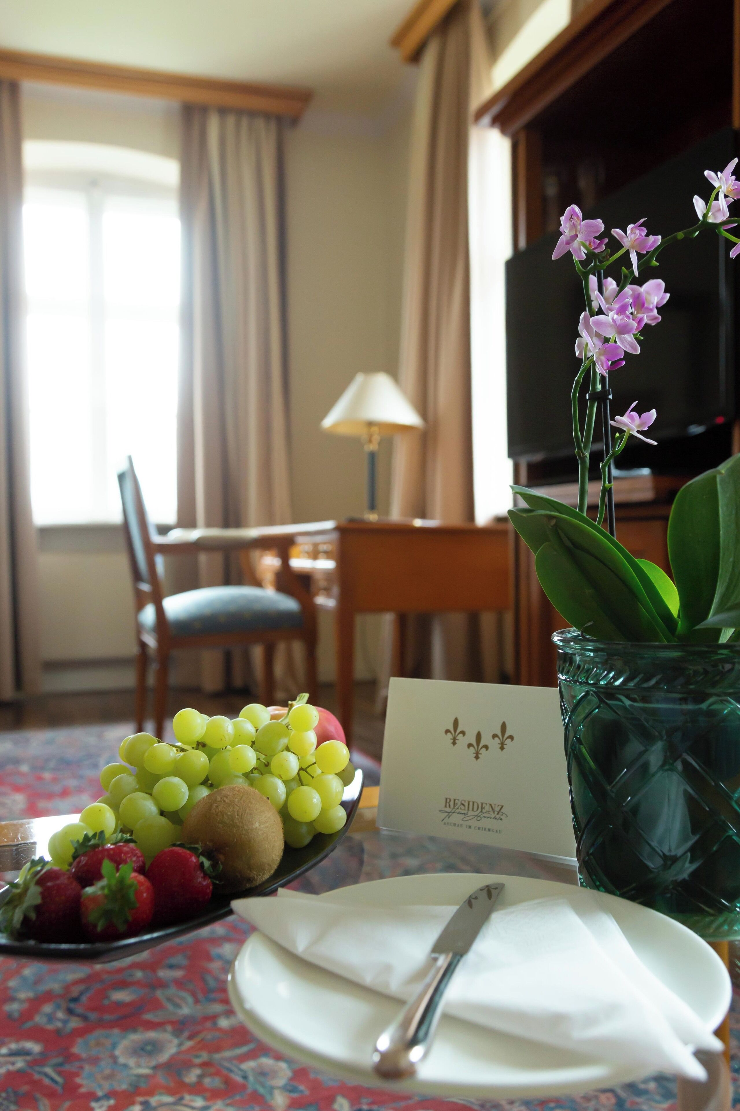 Habitación de hotel con una mesa de comedor para dos personas, un florero con flores y un frutero.