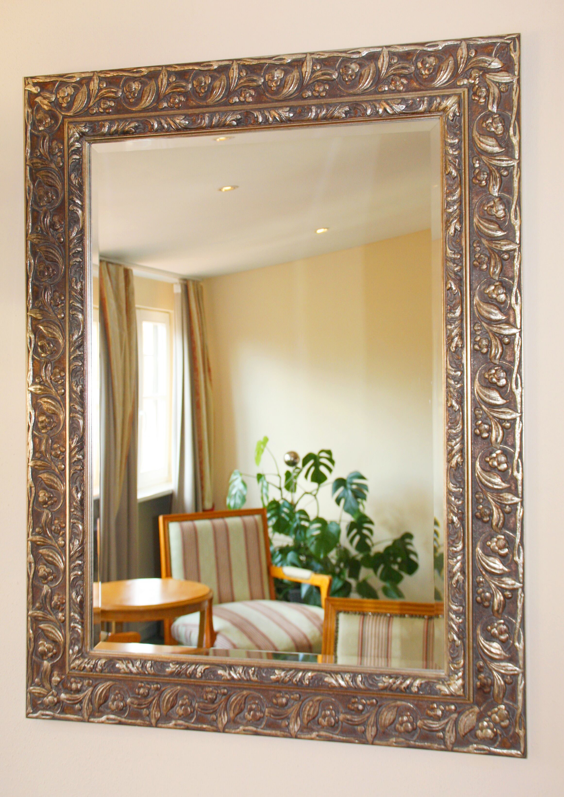 A mirror with an ornate frame reflecting a room with a chair, a small table, and a potted plant.